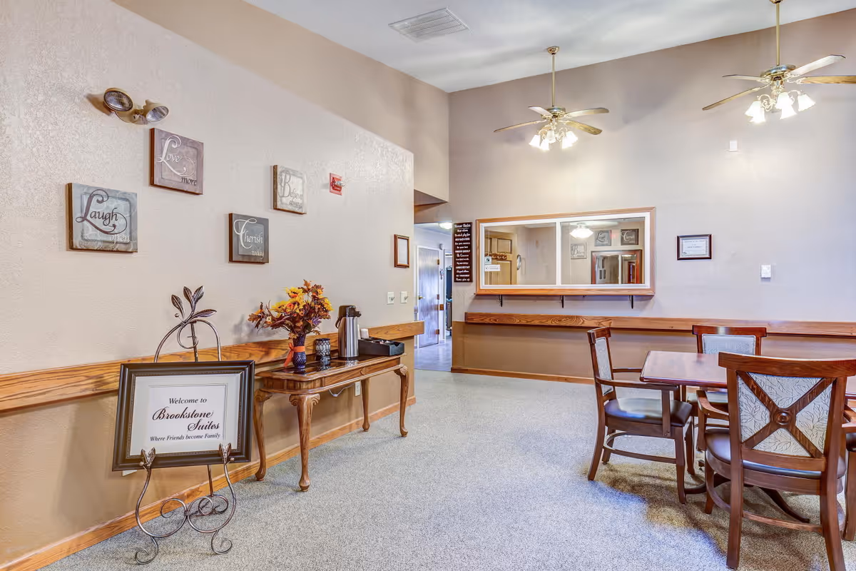 Interior view of a senior living facility common area with beige walls and carpeted floor. There is a wooden table with a coffee pot and a vase of flowers, a framed welcome sign for Brookstone Suites on a decorative stand, and several wall decorations with inspirational words. A wooden table with four chairs is positioned near a large window opening to another room. Two ceiling fans with lights hang from the ceiling.