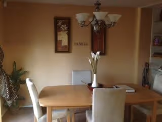 A dining area with a wooden table and four chairs, two of which are visible. A vase with white flowers is placed on the table. On the wall behind the table, there are two framed pictures and a decorative word. A chandelier with five lights hangs above the table. A plant is visible to the left side of the image.