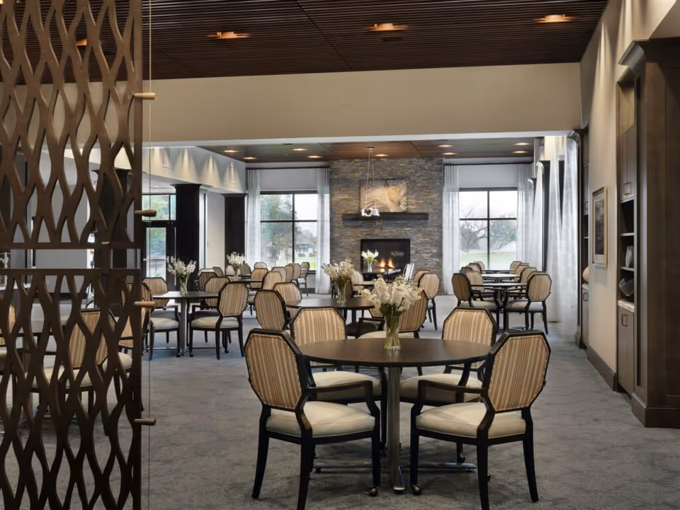 Spacious dining room with round tables and striped upholstered chairs, floral centerpieces, large windows, and a fireplace at the far wall.