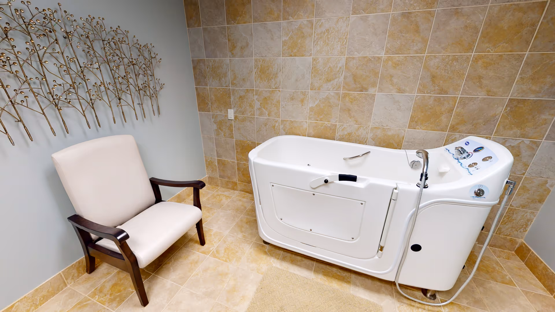 A room with beige tiled walls and floor featuring a white walk-in bathtub with control knobs and a handheld showerhead. Next to the bathtub is a cushioned armchair with dark wooden arms and legs. A decorative metal wall art piece resembling branches with small buds is mounted on the adjacent wall.