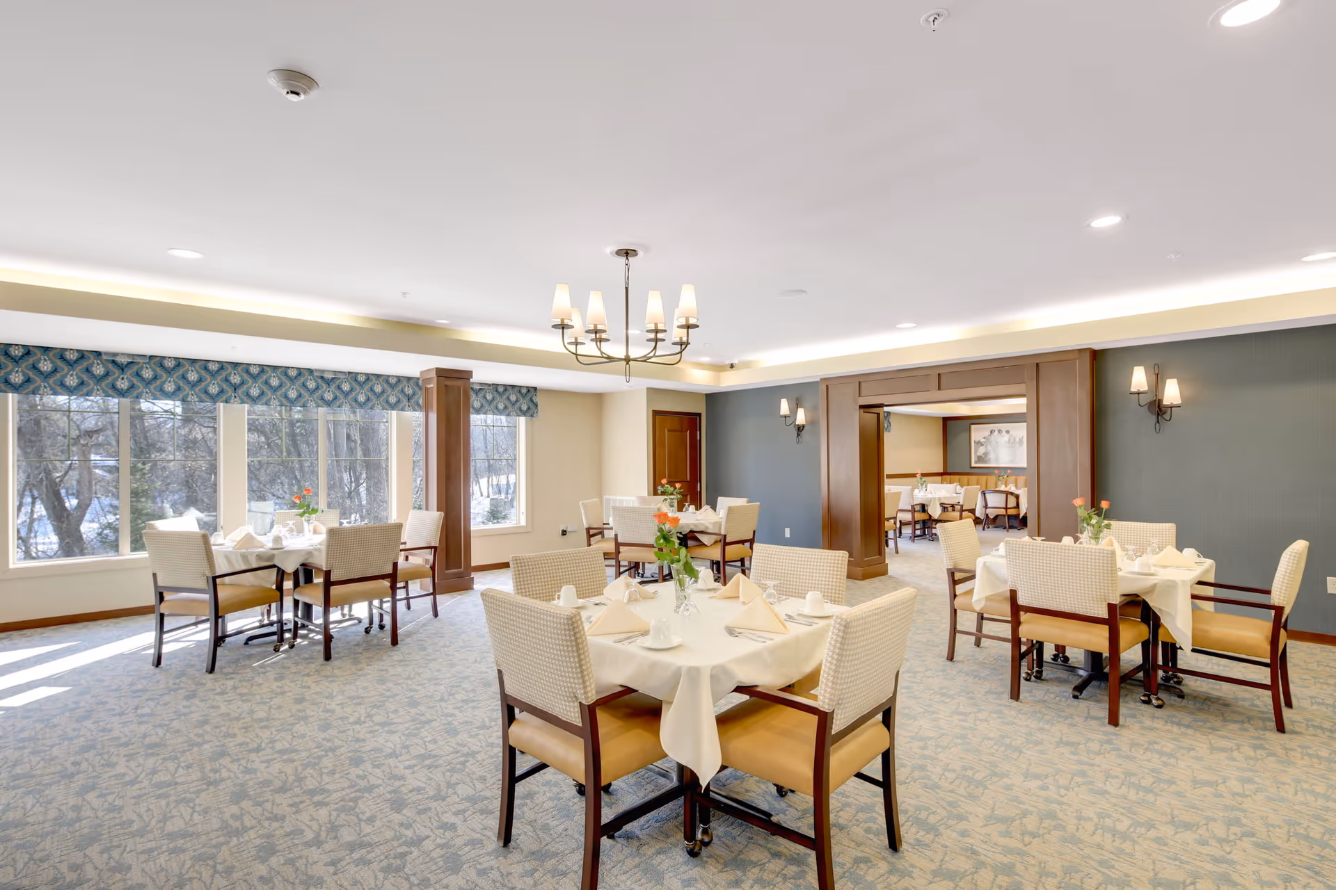 A bright and spacious dining room with several tables covered in white tablecloths, each set with napkins, cups, and small flower vases. The room has large windows letting in natural light, patterned carpet, and a chandelier hanging from the ceiling. There is an adjoining room visible through a wide doorway, also set up with dining tables and chairs.