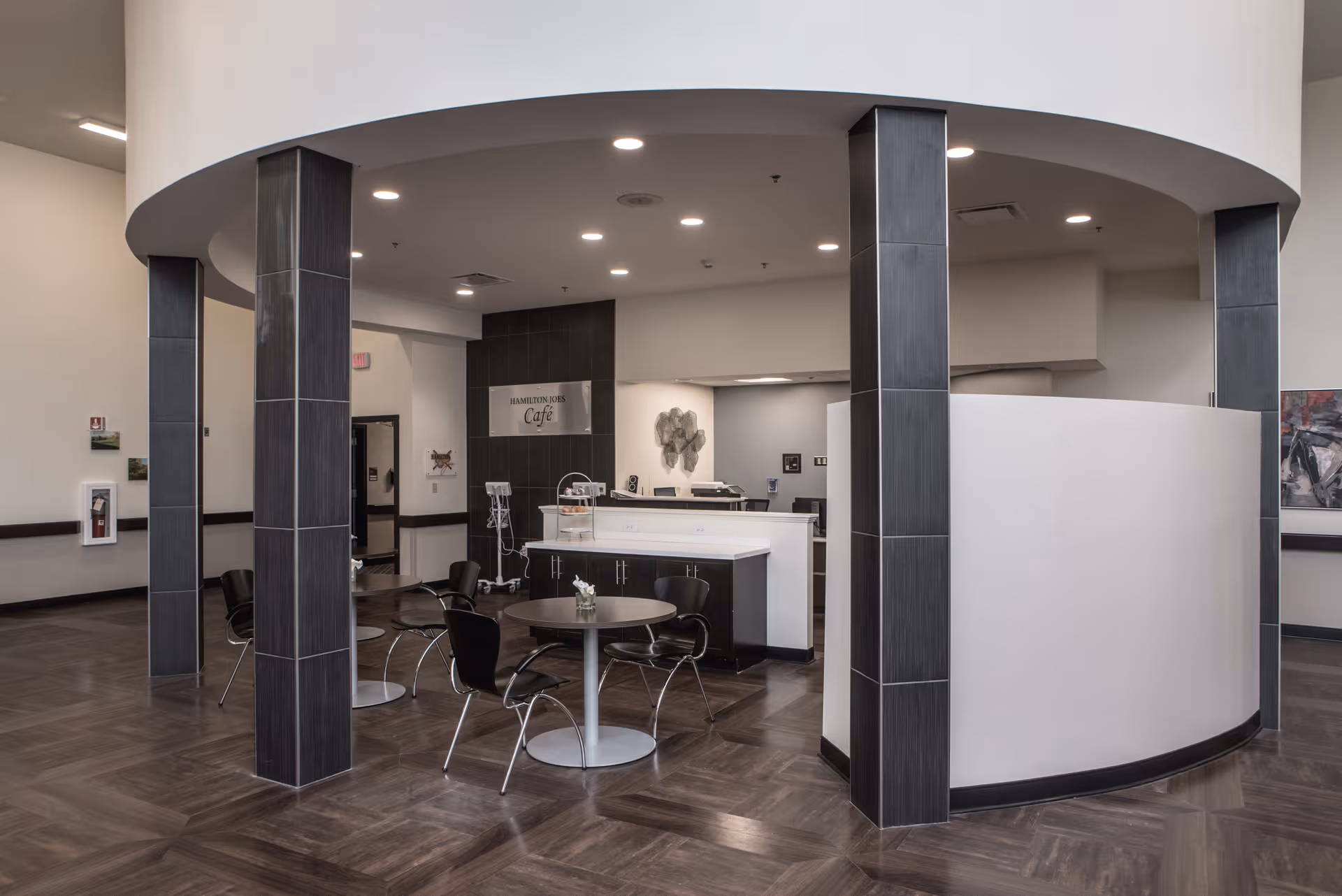 Interior view of a modern café area inside a senior living facility with round tables and black chairs. The space features dark tiled columns, wood-patterned flooring, and a white curved wall. A sign on the back wall reads 'Hamilton Jobs Café'.