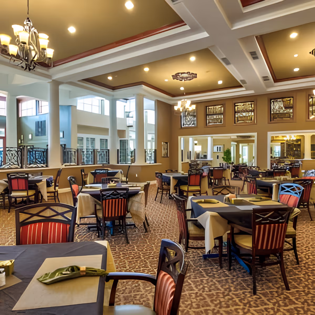 A spacious dining room with multiple tables covered with tablecloths and set with napkins. The room features a patterned carpet, large windows allowing natural light, decorative ceiling with recessed lighting, and chandeliers. Chairs with red and beige upholstery surround the tables, and mirrors adorn the walls.
