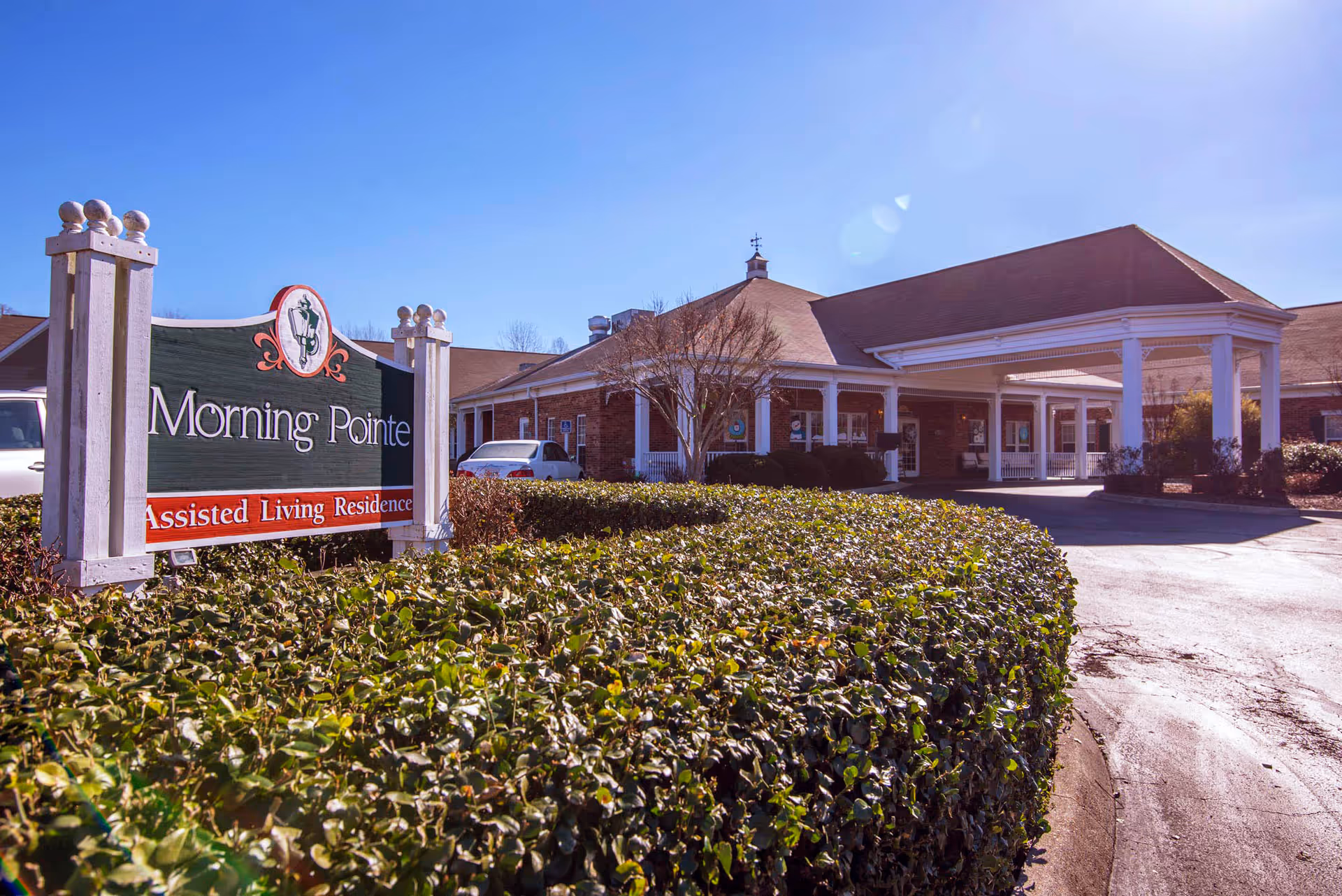 Exterior view of Morning Pointe Assisted Living Residence building with a clear blue sky, a green hedge in the foreground, and a sign displaying the facility's name.