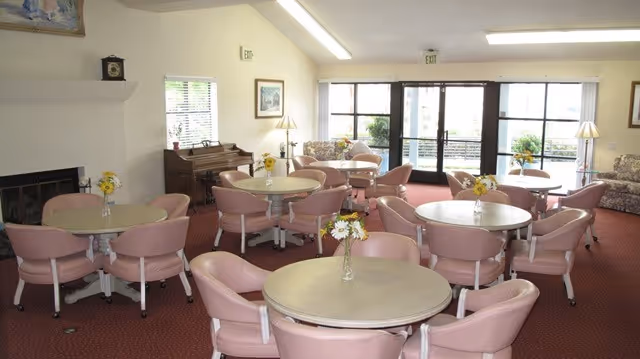 A senior living facility common area with multiple round tables surrounded by pink cushioned chairs. The room has a red carpet, a fireplace on the left, a piano against the wall, and large windows letting in natural light. There are floral arrangements on each table and a sofa near the windows.