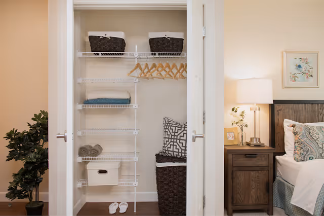 Open closet with white wire shelves holding folded towels, baskets, and wooden hangers. To the right is a bedside table with a lamp, a small plant, and a framed photo next to a bed with patterned pillows and a white blanket. A potted plant is visible on the left side of the closet.