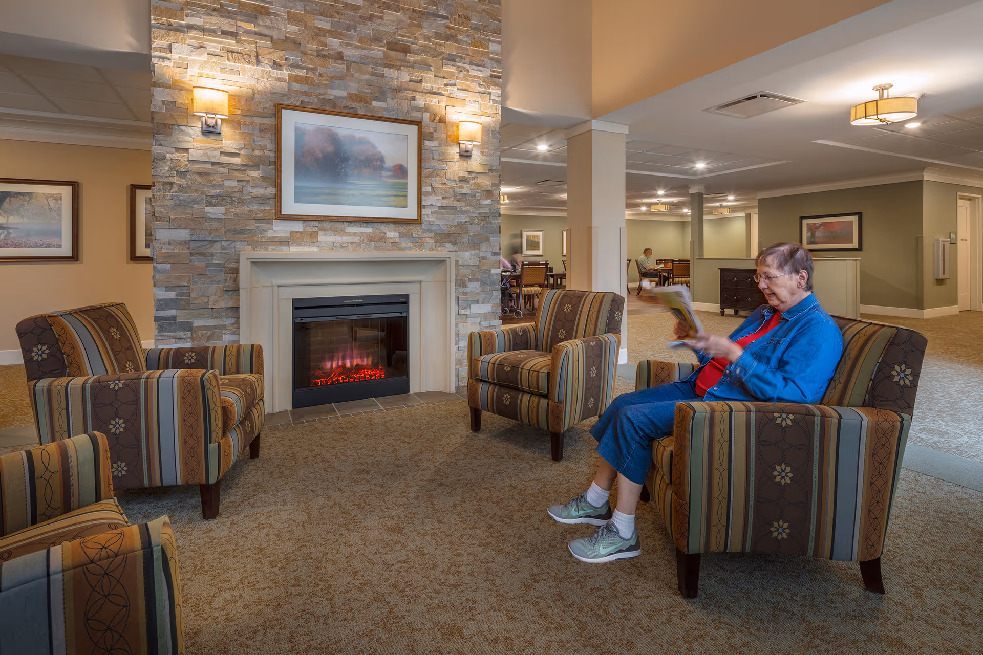 A cozy common area in a senior living facility with a stone fireplace, three striped armchairs arranged around it, and an elderly woman sitting in one chair reading a magazine. The room has warm lighting, framed artwork on the walls, and a carpeted floor.