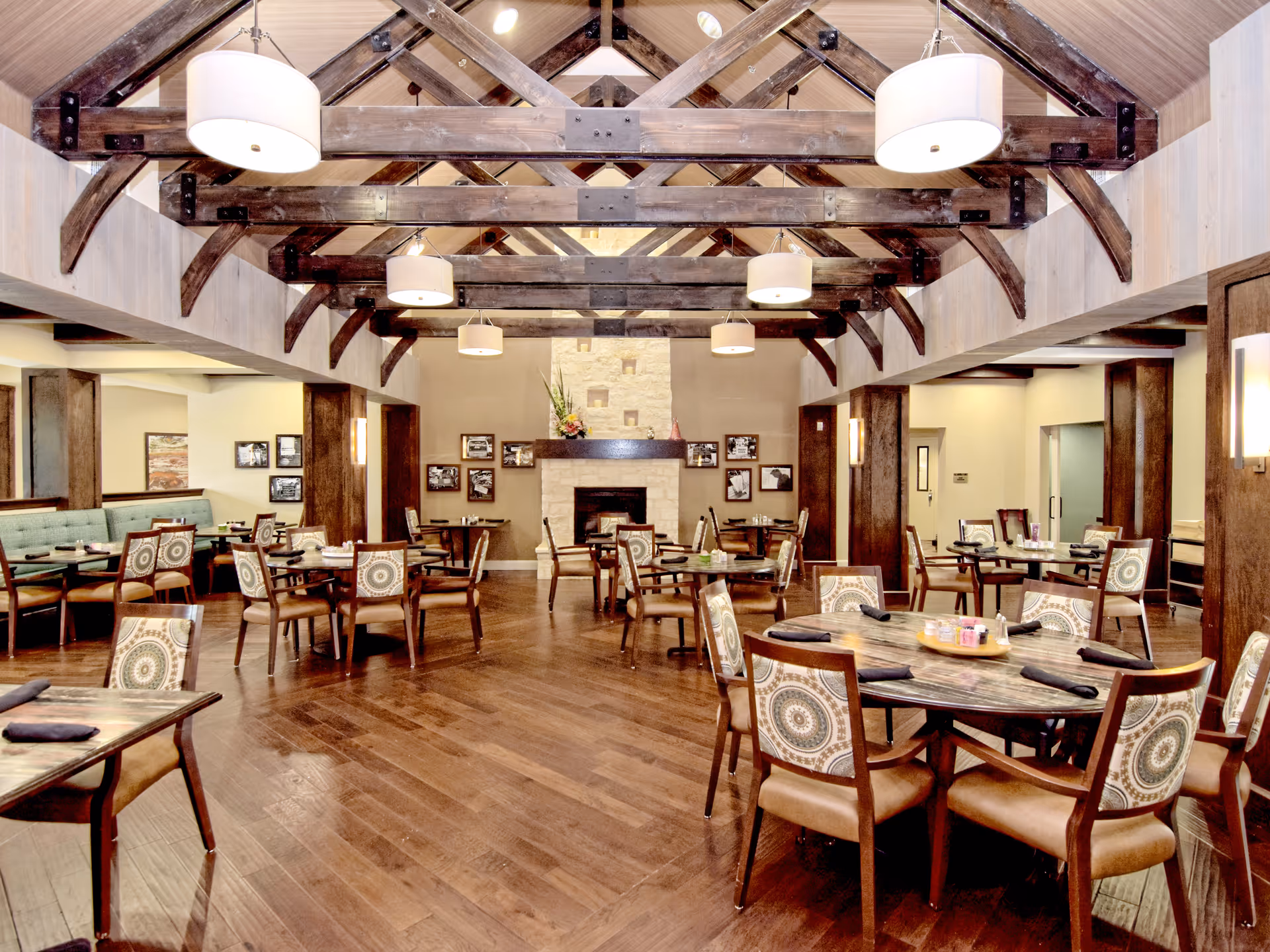 A spacious dining room with wooden floors, multiple round and rectangular tables with patterned chairs, a stone fireplace at the far end, and exposed wooden beams on the ceiling with hanging light fixtures.