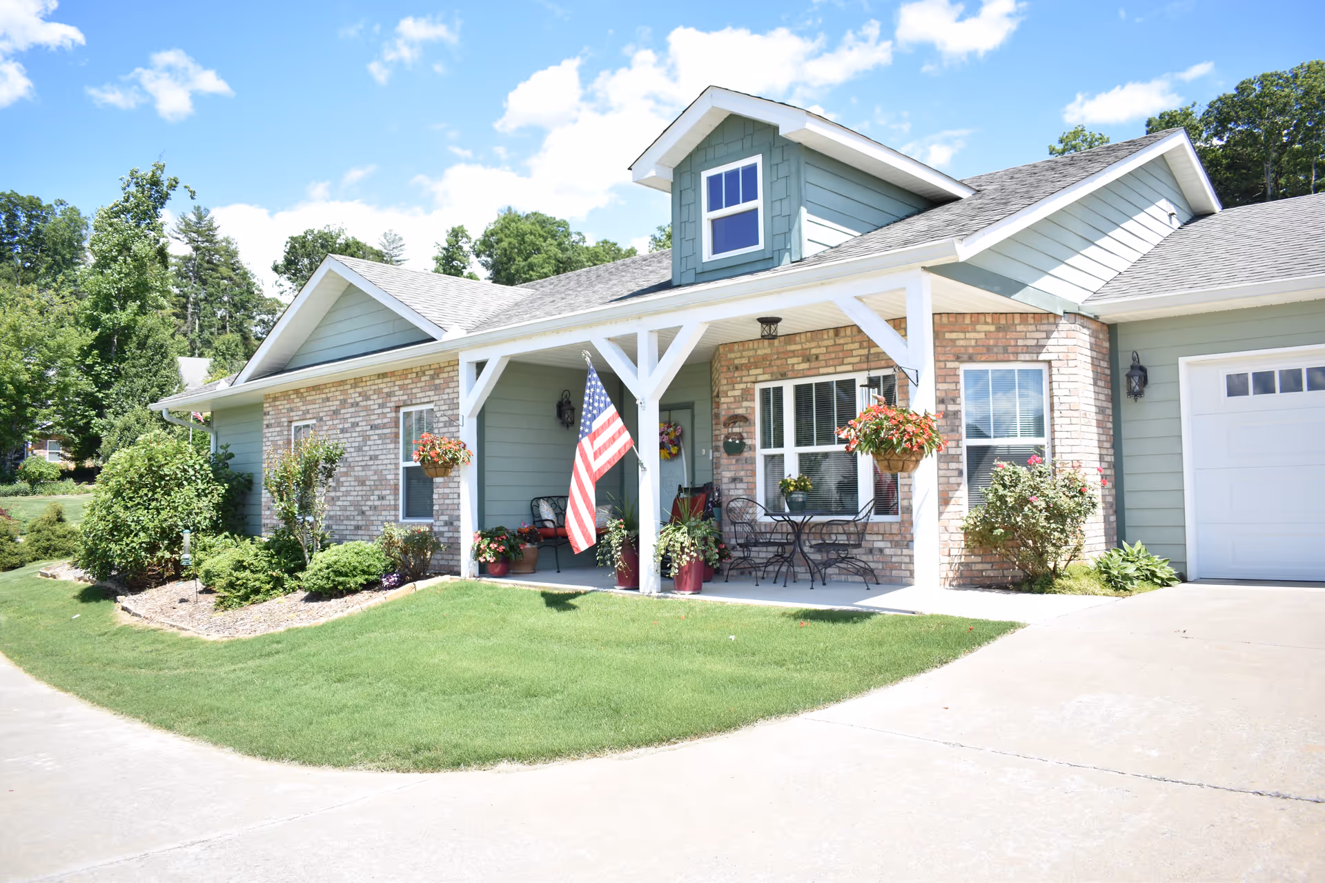 Front exterior of a single-story house with a covered porch, American flag, potted plants, and attached garage.