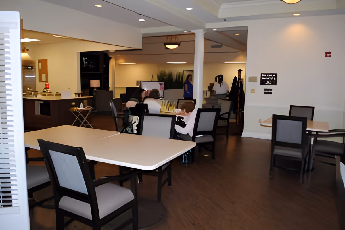 Interior view of a senior living facility common area with several tables and chairs arranged for seating. Some residents are seated and engaged in activities. The room has wooden flooring, white walls, and ceiling lights. There is a counter area in the background with some items on it.