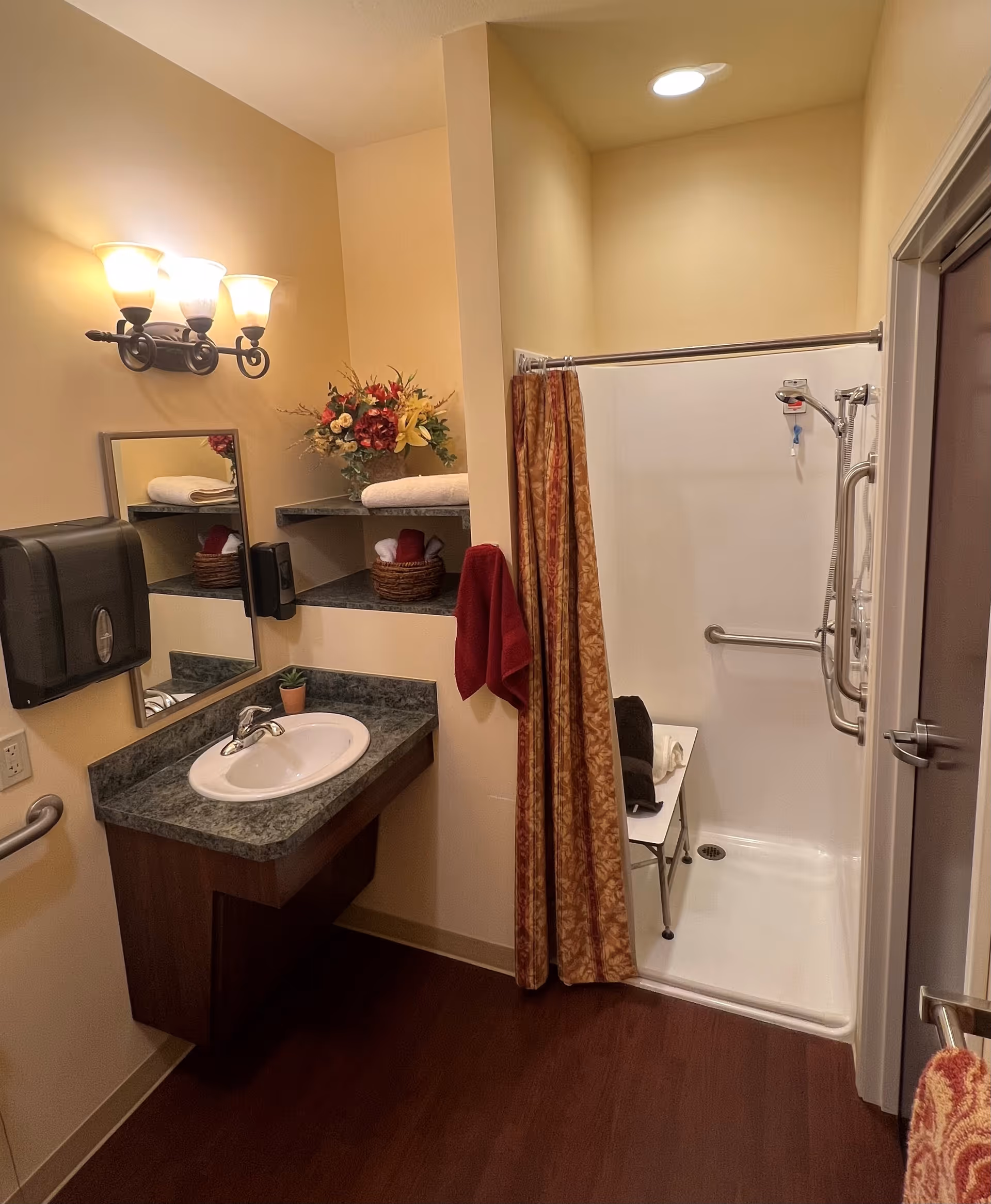 Accessible bathroom with a sink and mirror, shelving with towels and flowers, and a roll-in shower with grab bars and a shower curtain.