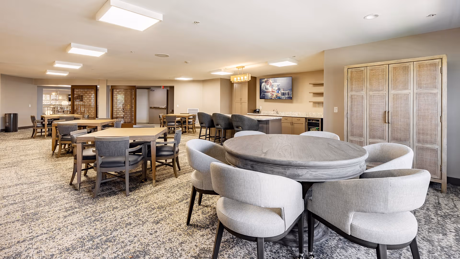 A spacious and well-lit dining area with multiple wooden tables and chairs arranged neatly. In the foreground, there is a round wooden table surrounded by six cushioned chairs. The room features a carpeted floor, modern ceiling lights, a small kitchenette area with cabinets, a countertop, and a wall-mounted TV displaying an image. Decorative wooden doors and neutral-colored walls complete the setting.