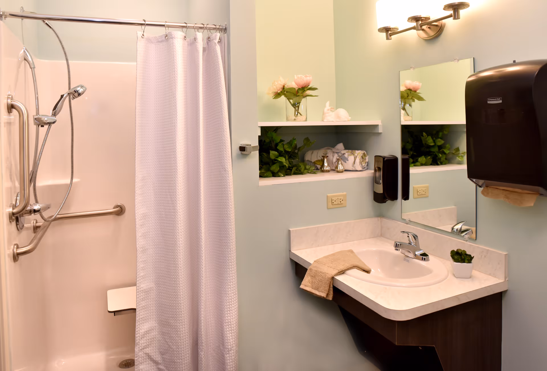 A clean accessible bathroom with a shower and grab bars, a sink with mirror, and decorative shelving with plants.