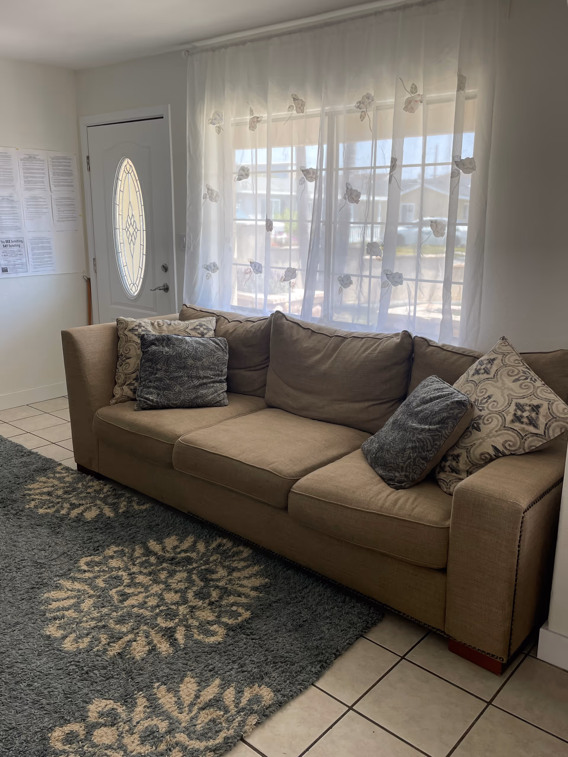 A beige sofa with patterned throw pillows sits on a gray rug with floral designs in a room with tiled floors. Behind the sofa is a window covered with sheer white curtains decorated with floral embroidery. To the left of the sofa is a white door with an oval glass window, and some papers are posted on the adjacent wall.