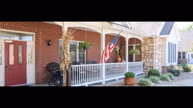 Front porch of a brick and stone building with a red door, white railing, hanging plants, an American flag, and a wheelchair parked near the entrance.