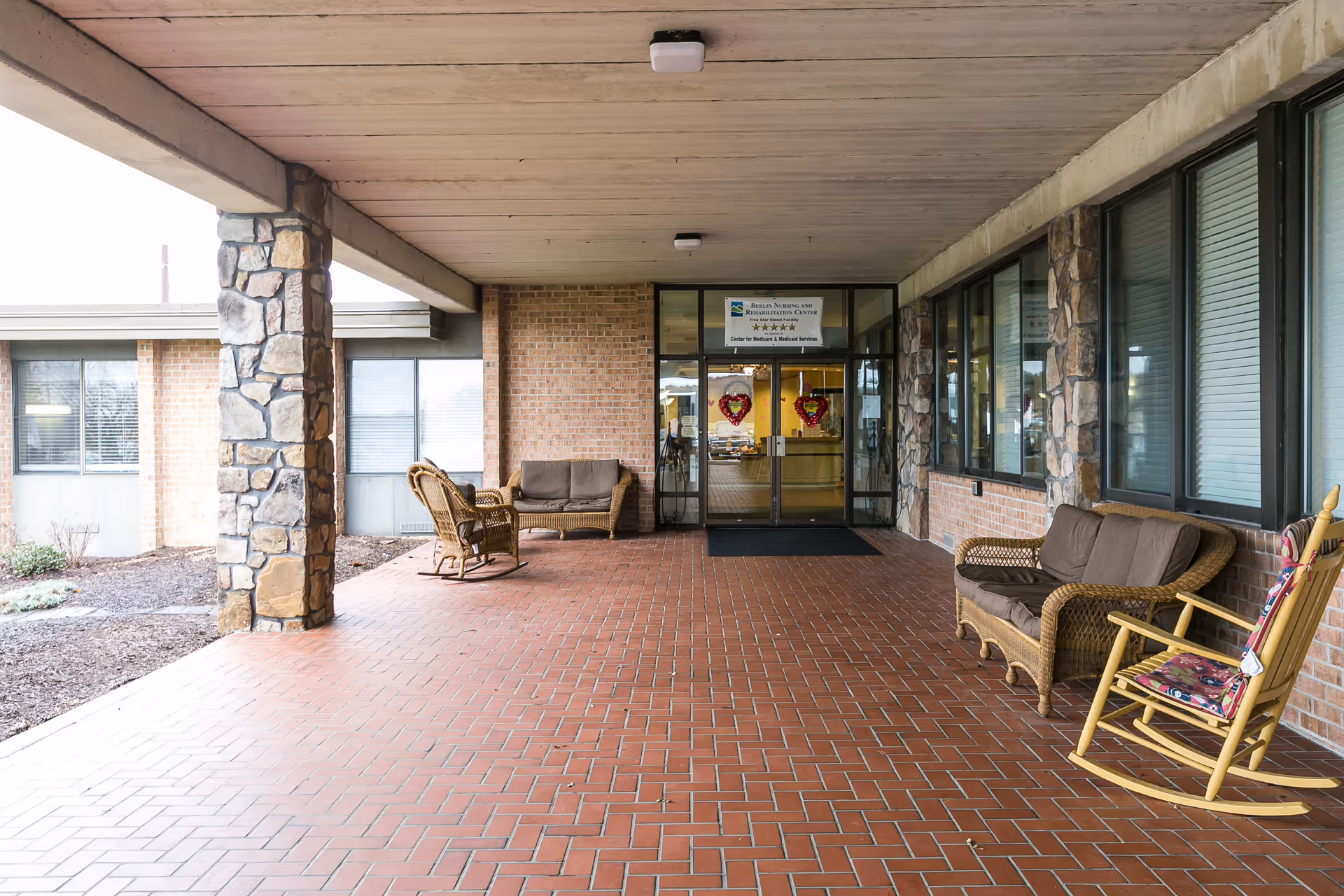 Covered entrance area of Berlin Nursing and Rehabilitation facility with brick flooring, stone pillars, wicker seating including chairs and a loveseat, and a yellow rocking chair with a floral cushion. Glass double doors with heart-shaped decorations lead inside.