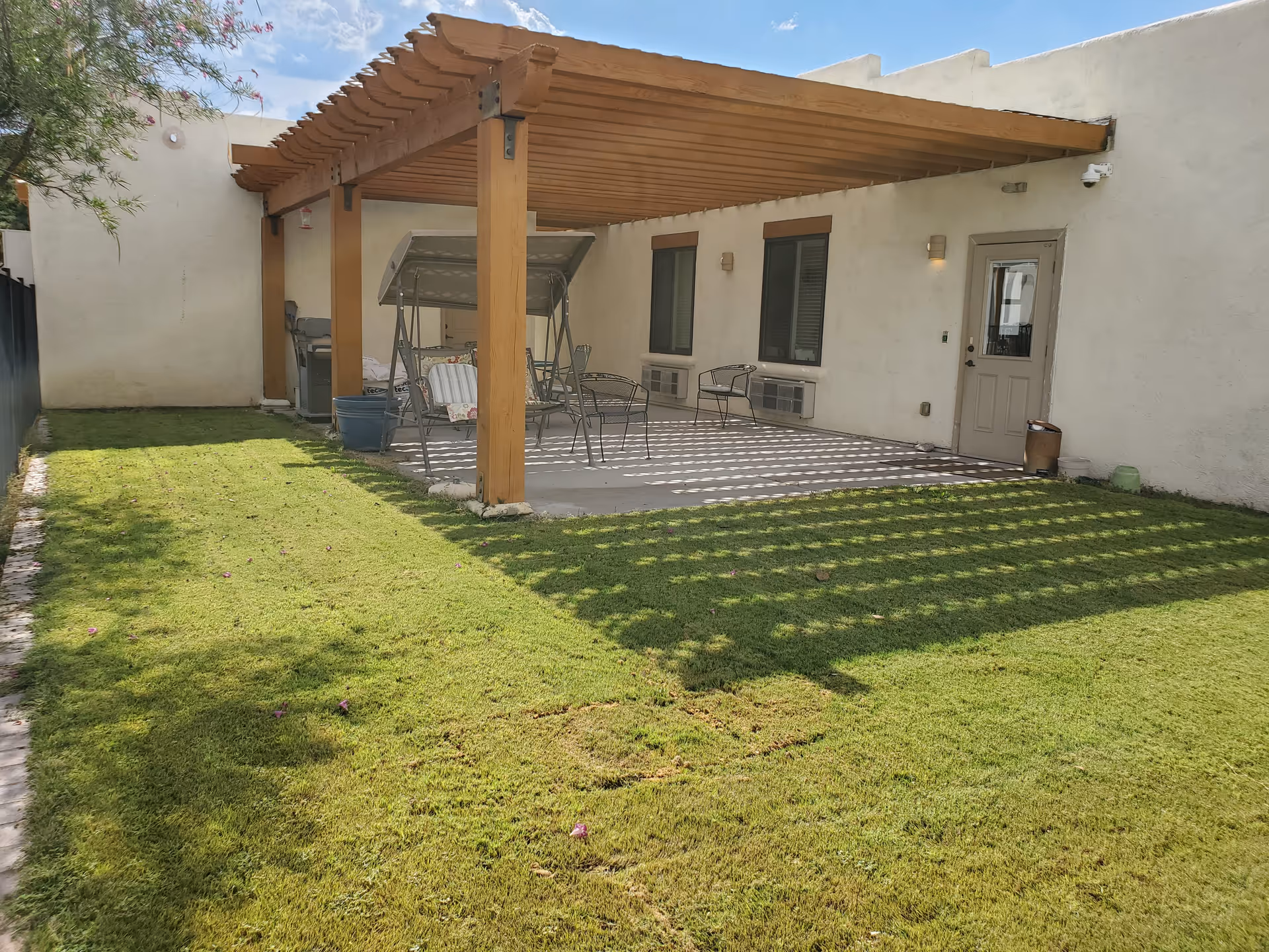 A grassy backyard with a wooden pergola-covered patio, metal chairs and a swing next to a stucco building.