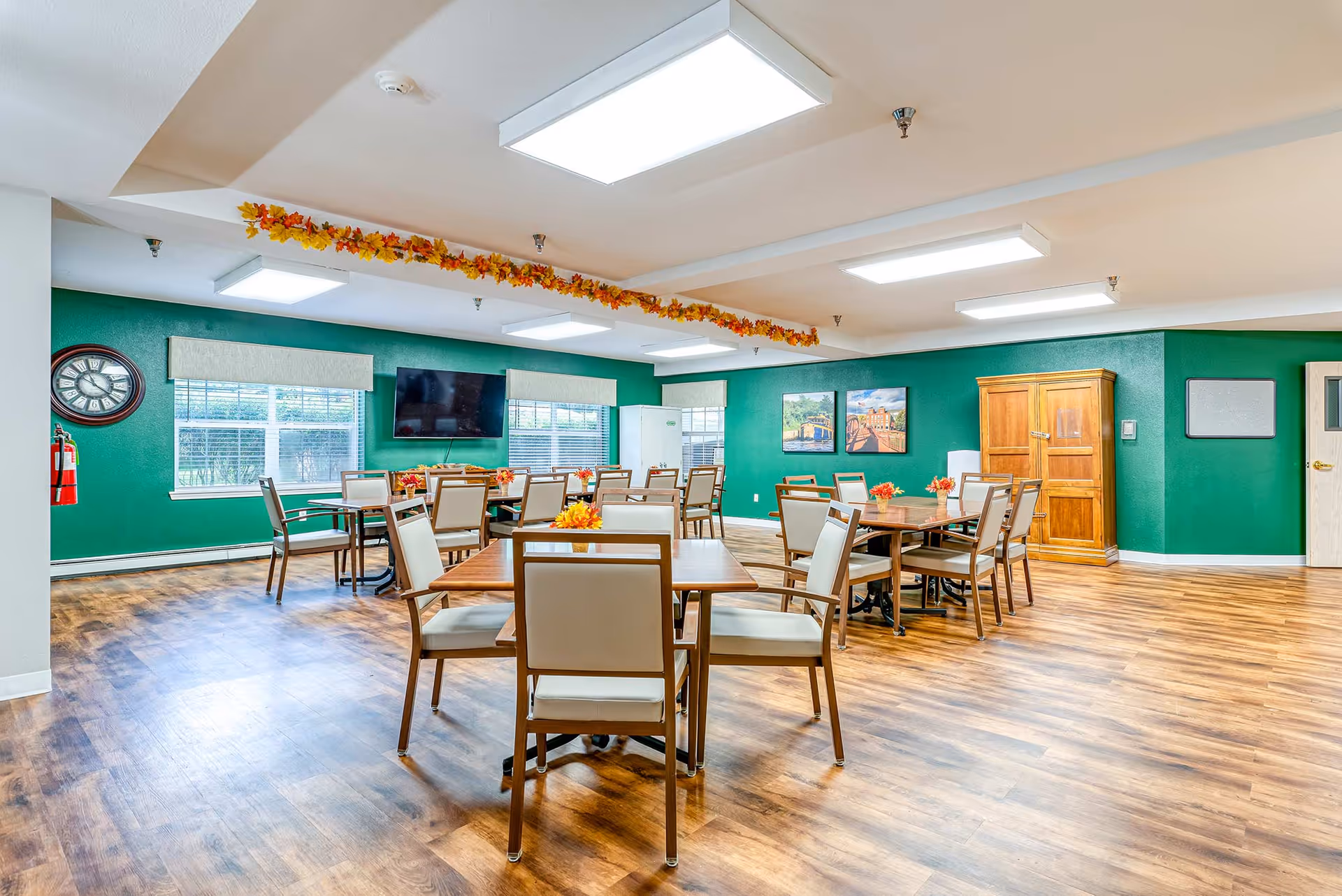 A bright and spacious dining room with multiple wooden tables and chairs arranged neatly. The walls are painted green and decorated with framed pictures and a large clock. There is a flat-screen TV mounted on the wall, windows with blinds, and a wooden cabinet. The ceiling has rectangular fluorescent lights and a garland of autumn leaves hanging across.