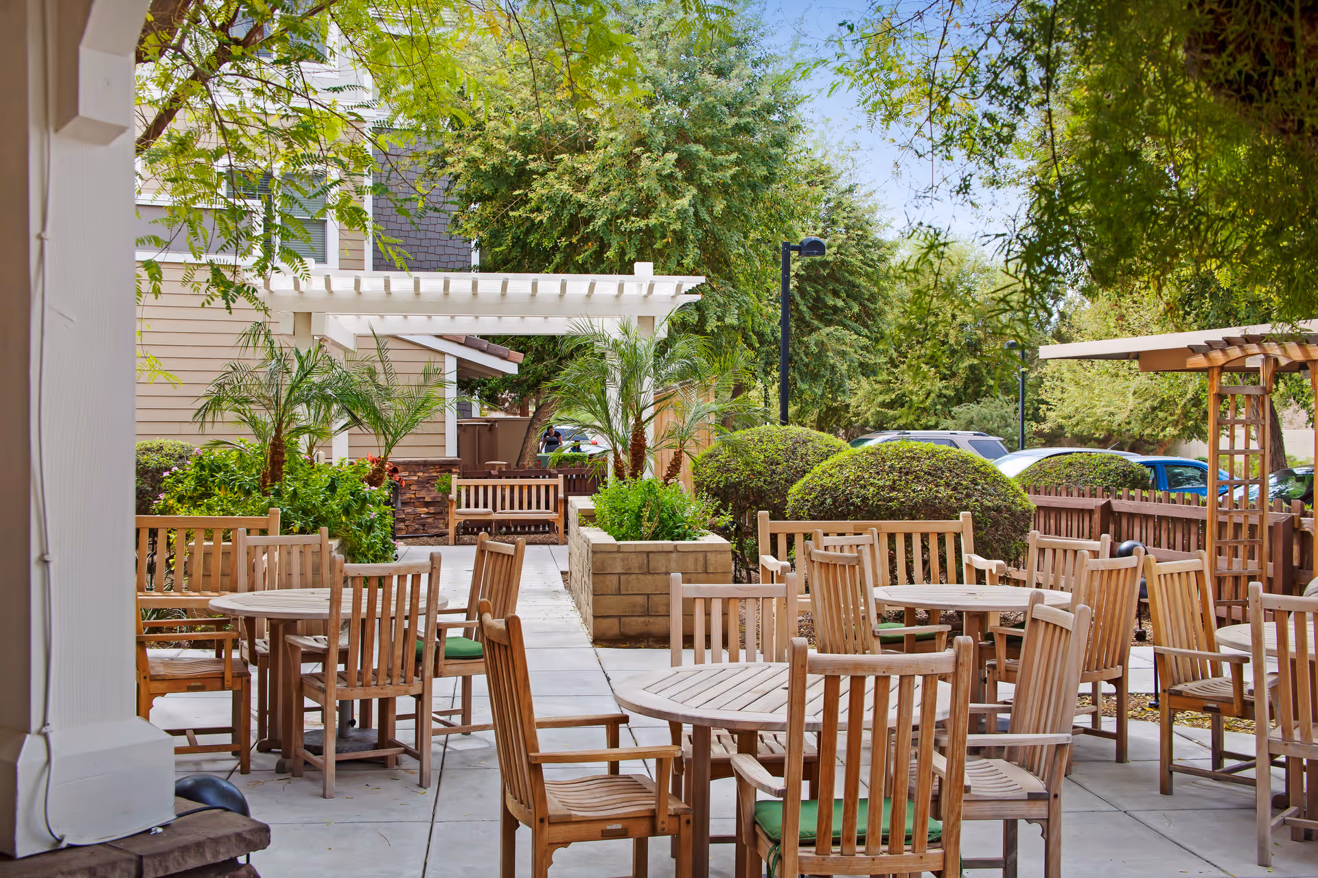 Outdoor seating area with multiple wooden tables and chairs with green cushions, surrounded by lush greenery, bushes, palm plants, and trees. There is a white pergola and a wooden trellis, with a building and parked cars visible in the background under a clear sky.