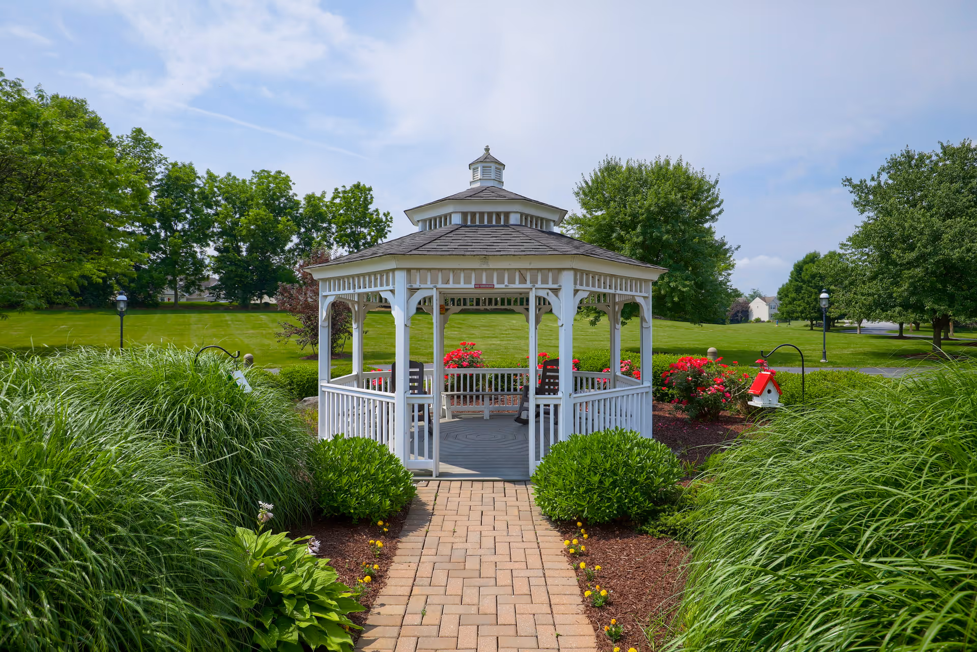 A white wooden gazebo with benches inside, situated in a well-maintained garden area with green bushes, flowering plants, and a paved walkway leading to it. The sky is partly cloudy and trees surround the garden.