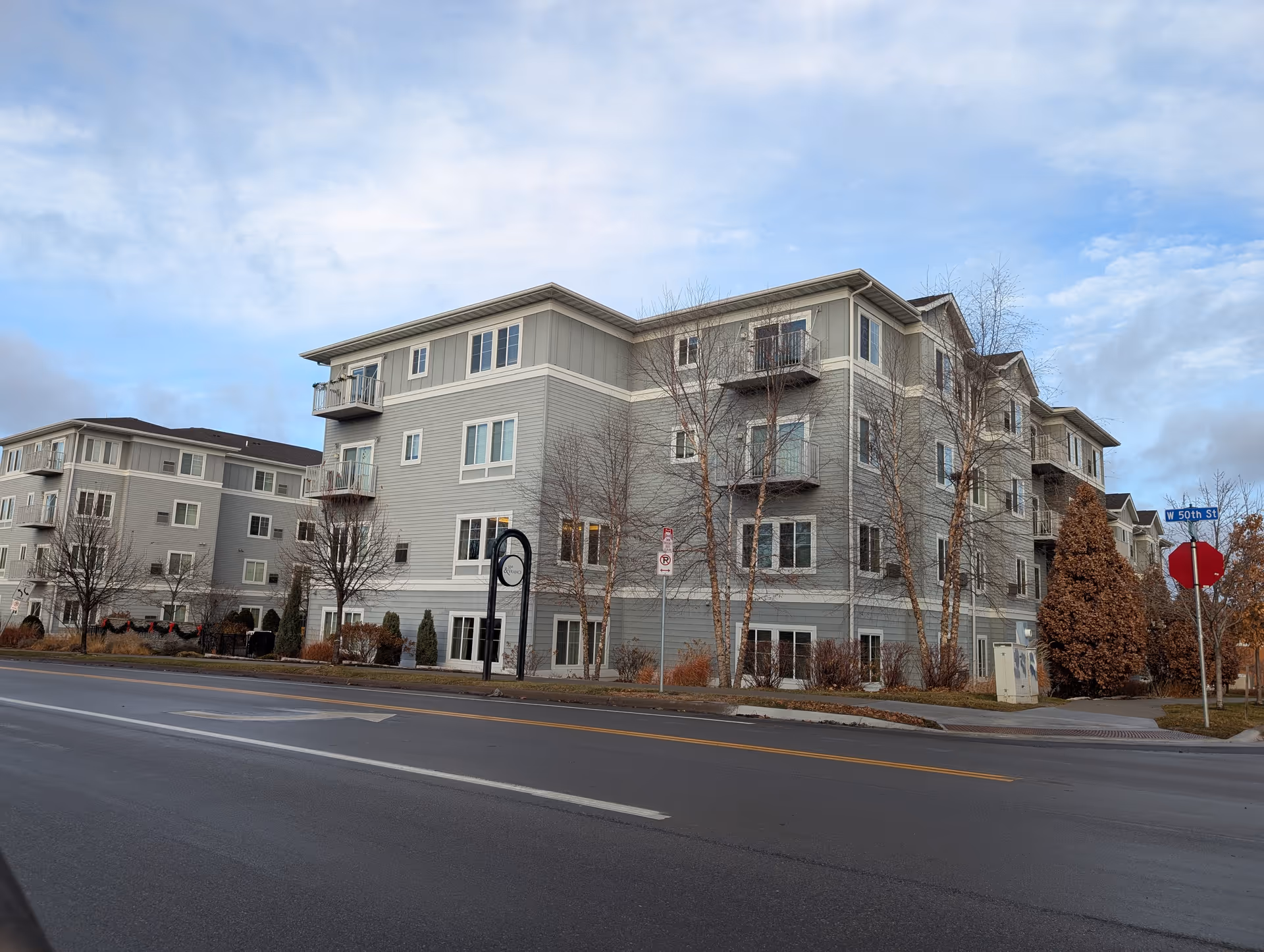 Four-story gray apartment-style senior living building with balconies on a street corner, bare trees, and a stop sign.
