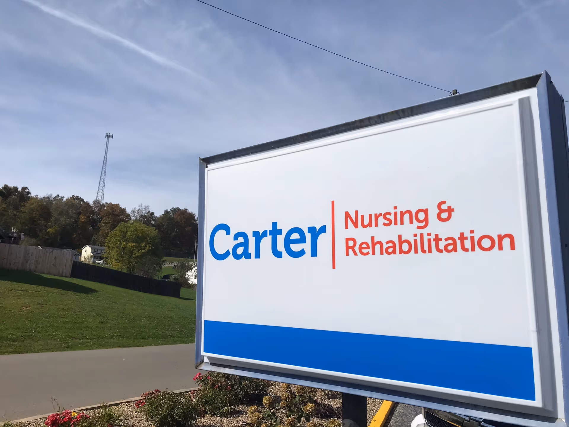 Outdoor view of a large sign for Carter Nursing & Rehabilitation with a grassy area, trees, and a communication tower in the background under a partly cloudy sky.