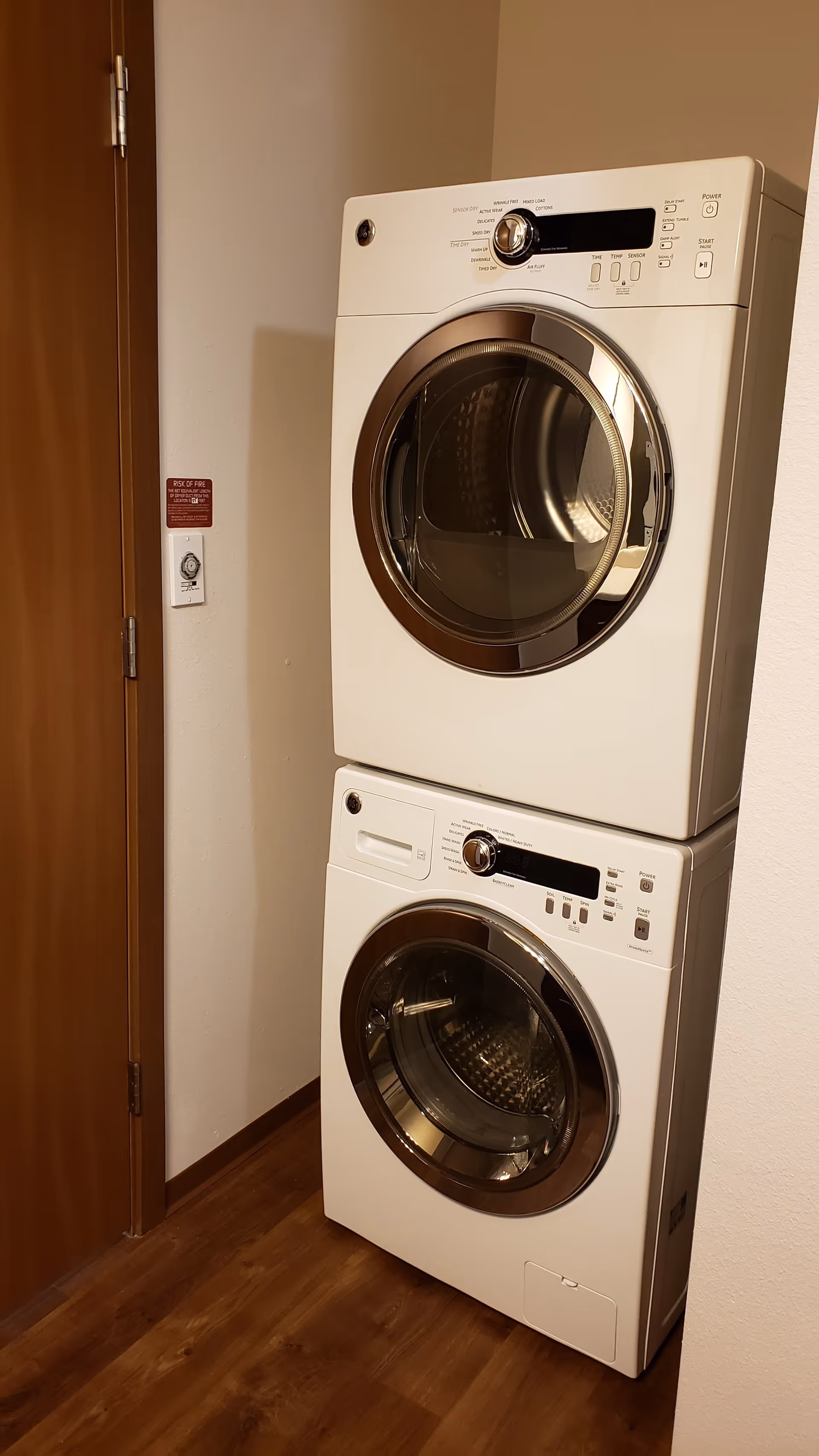 Stacked white front-loading washer and dryer units in a small laundry area with wooden floor and a wooden door to the left.