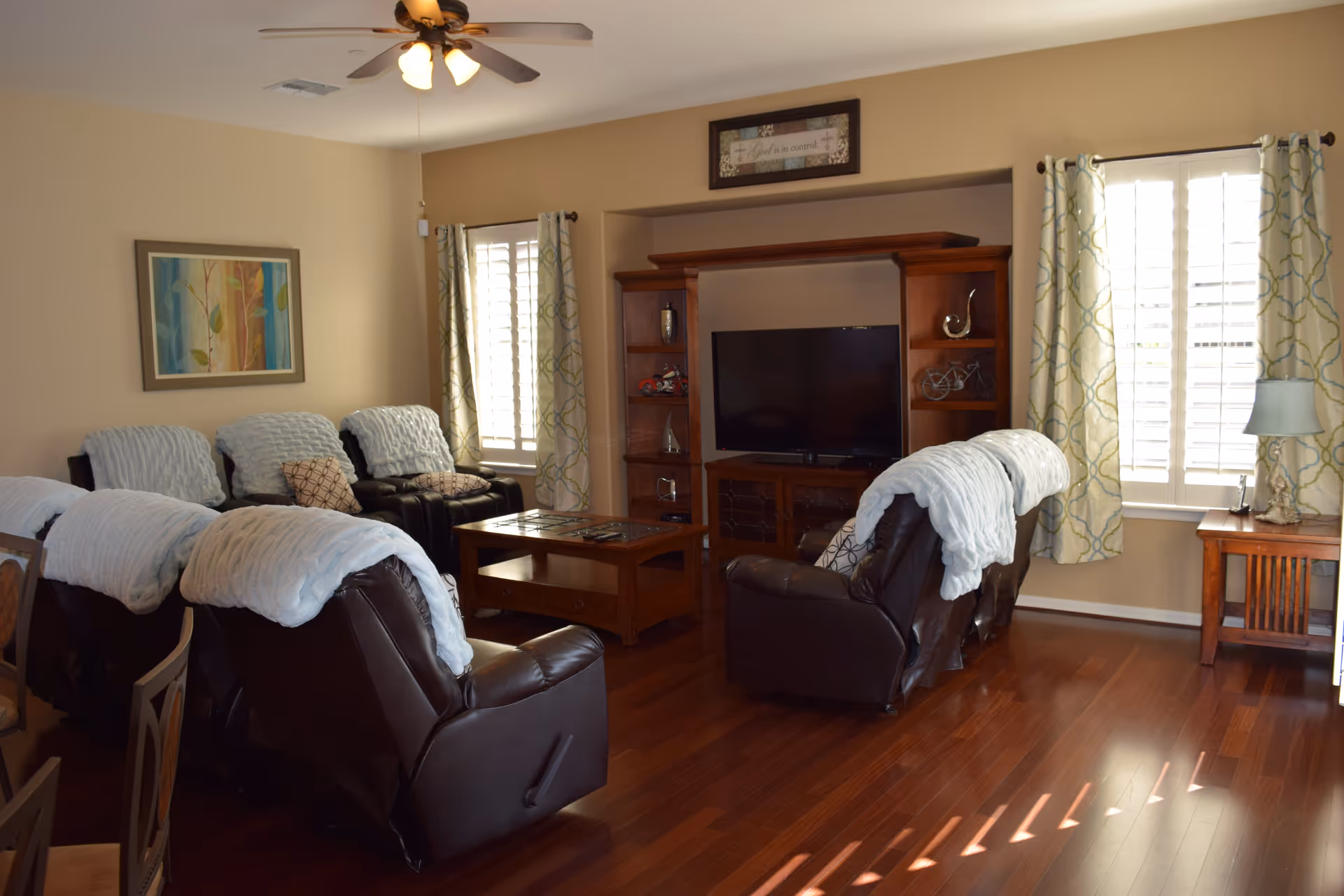 Sunlit living room with several leather recliners draped in white throws facing a wooden entertainment center and TV.