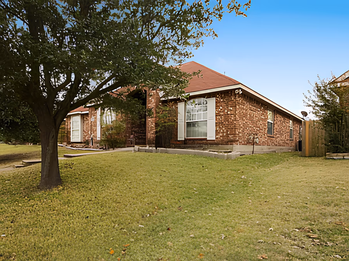 Exterior view of a single-story brick building with a red roof, white window shutters, and a large tree in the grassy front yard under a clear blue sky.