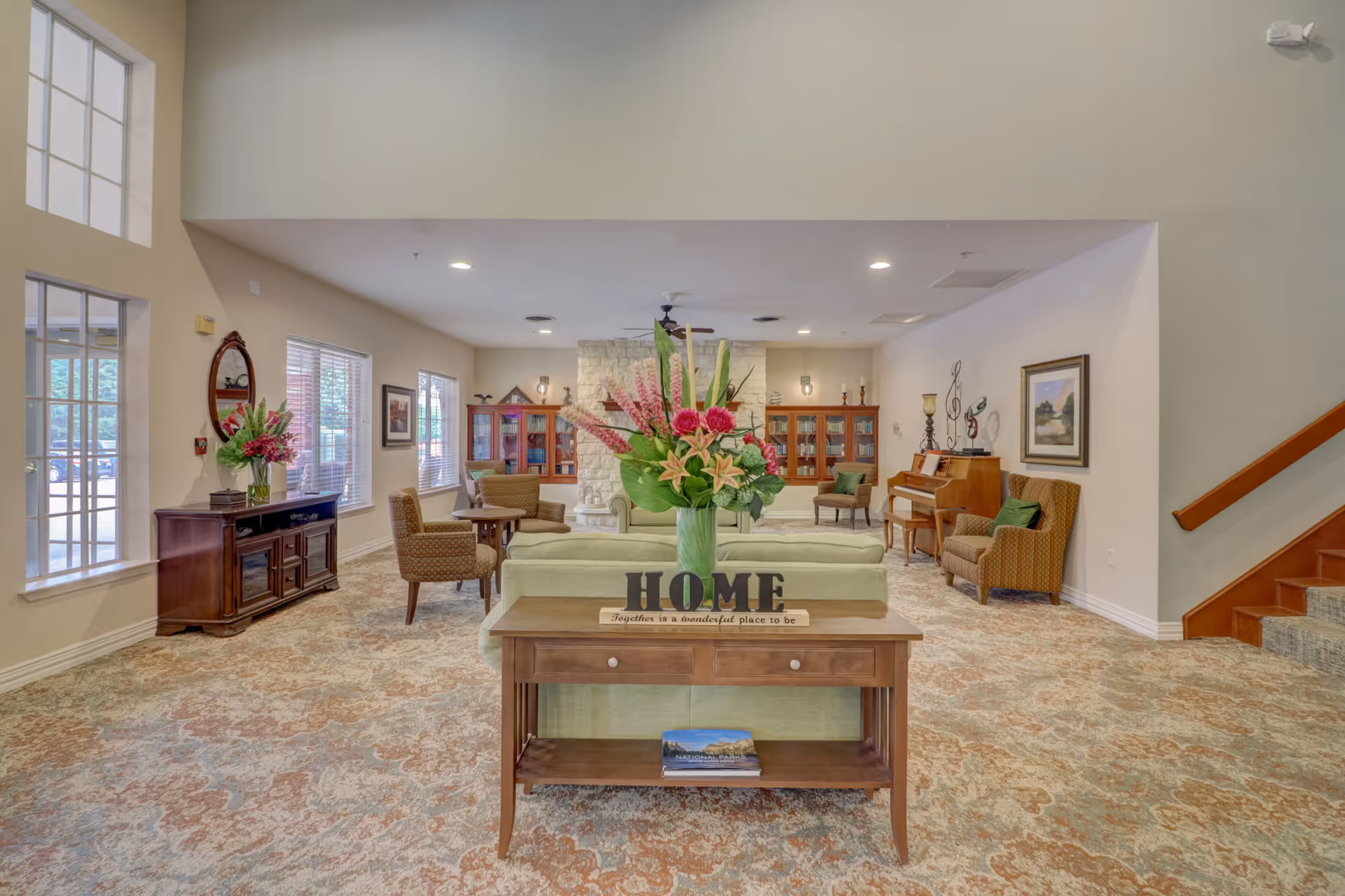 A spacious and well-lit living room area in a senior living facility with a patterned carpet, light green sofas, armchairs, a wooden table with a flower vase and a decorative 'HOME' sign, large windows, a stone fireplace, wooden bookshelves, a piano, and framed artwork on the walls.