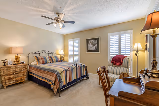 A cozy bedroom with a metal frame bed dressed in striped bedding. There are two windows with white shutters, a wooden nightstand with a lamp and decorative items, a striped armchair with a red throw blanket, and a wooden desk with a lamp and chair. The walls are painted beige and the room is carpeted.