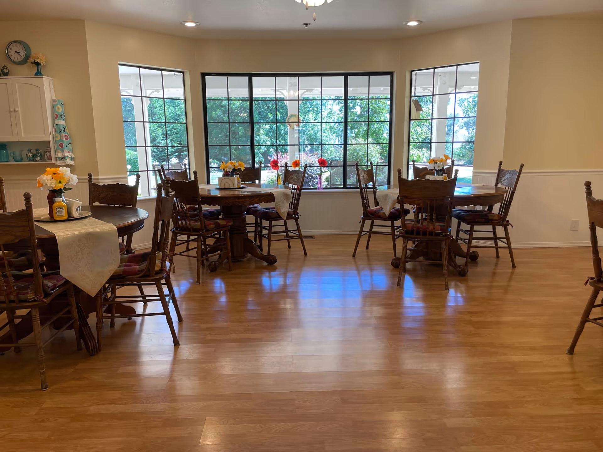 Bright dining room with several round wooden tables and chairs, floral centerpieces, and a large bay window overlooking greenery.