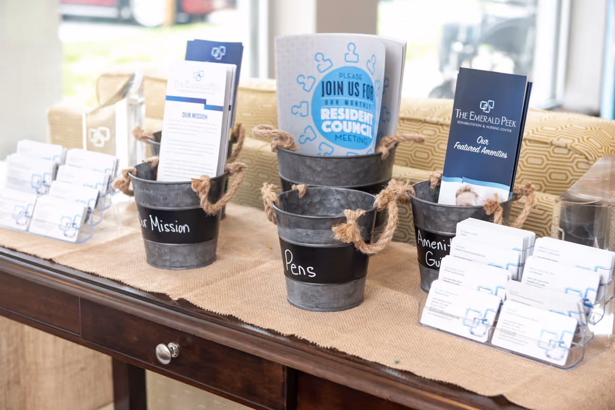 A wooden table with informational brochures and business cards displayed in metal buckets and clear holders. The buckets are labeled 'Our Mission,' 'Pens,' and 'Amenities Guide.' The brochures include information about The Emerald Peek Rehabilitation & Nursing Center and a flyer inviting residents to a monthly Resident Council meeting. The background shows a patterned couch and a window with natural light.