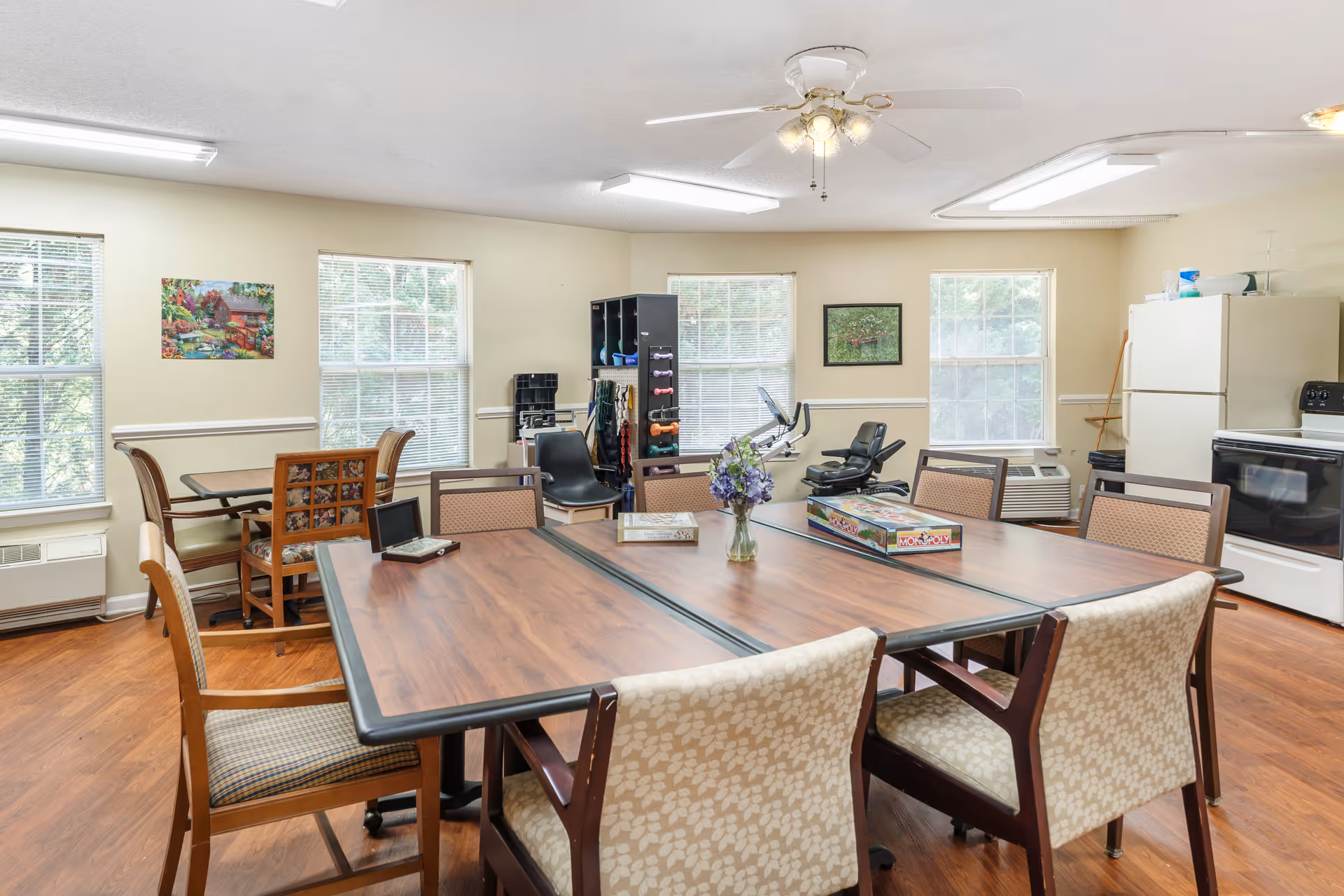 Bright communal dining/activity room with a large table surrounded by chairs, windows, a small kitchen area, and exercise equipment in the background.
