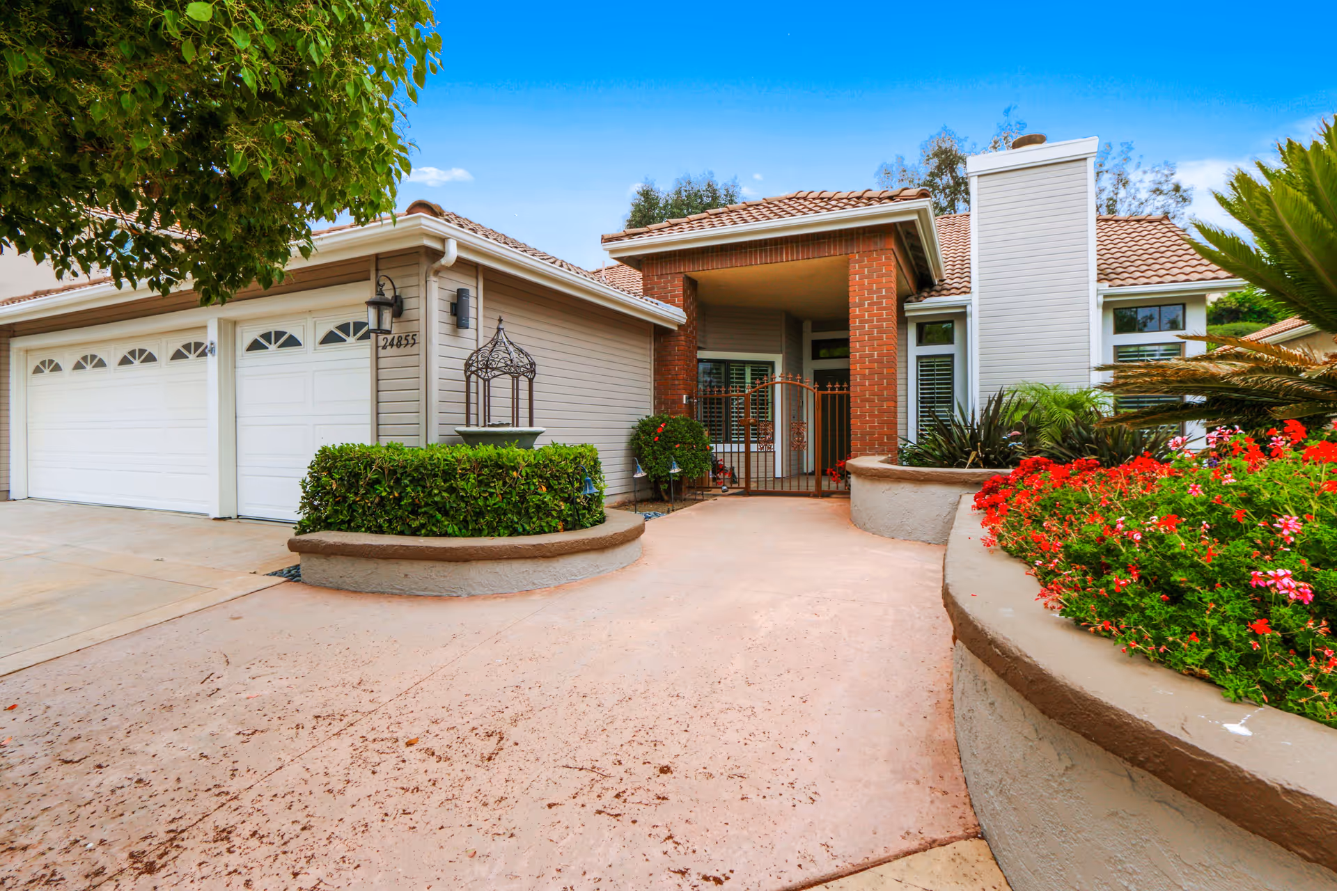 Front exterior view of a single-story residential building with a tiled roof, a three-car garage with white doors, a gated entrance, and landscaped garden beds with green shrubs and red flowers under a clear blue sky.