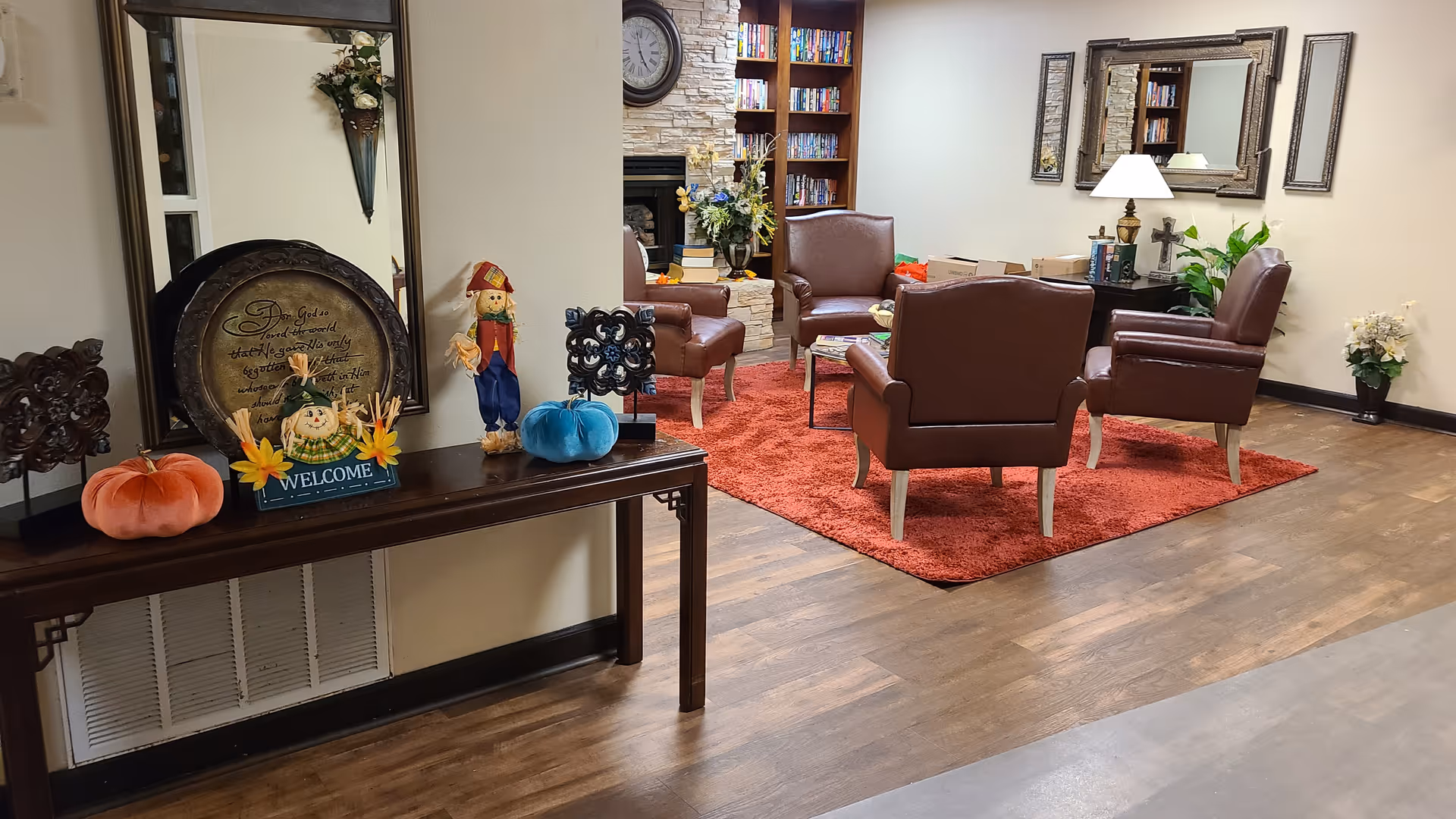 A cozy sitting area in a retirement center with four brown leather chairs arranged around a small table on a red rug. Behind the chairs is a stone fireplace with a clock above it and a bookshelf filled with books. To the left, a wooden console table holds autumn-themed decorations including fabric pumpkins, a scarecrow figurine, a welcome sign, and a decorative plate with an inspirational quote. The walls are light-colored and decorated with mirrors and framed art, and there are plants placed around the room.