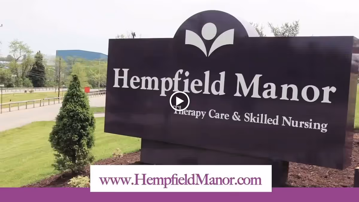 Outdoor view of a large dark purple sign for Hempfield Manor Therapy Care & Skilled Nursing, with a small tree and green grass around it and a road in the background.
