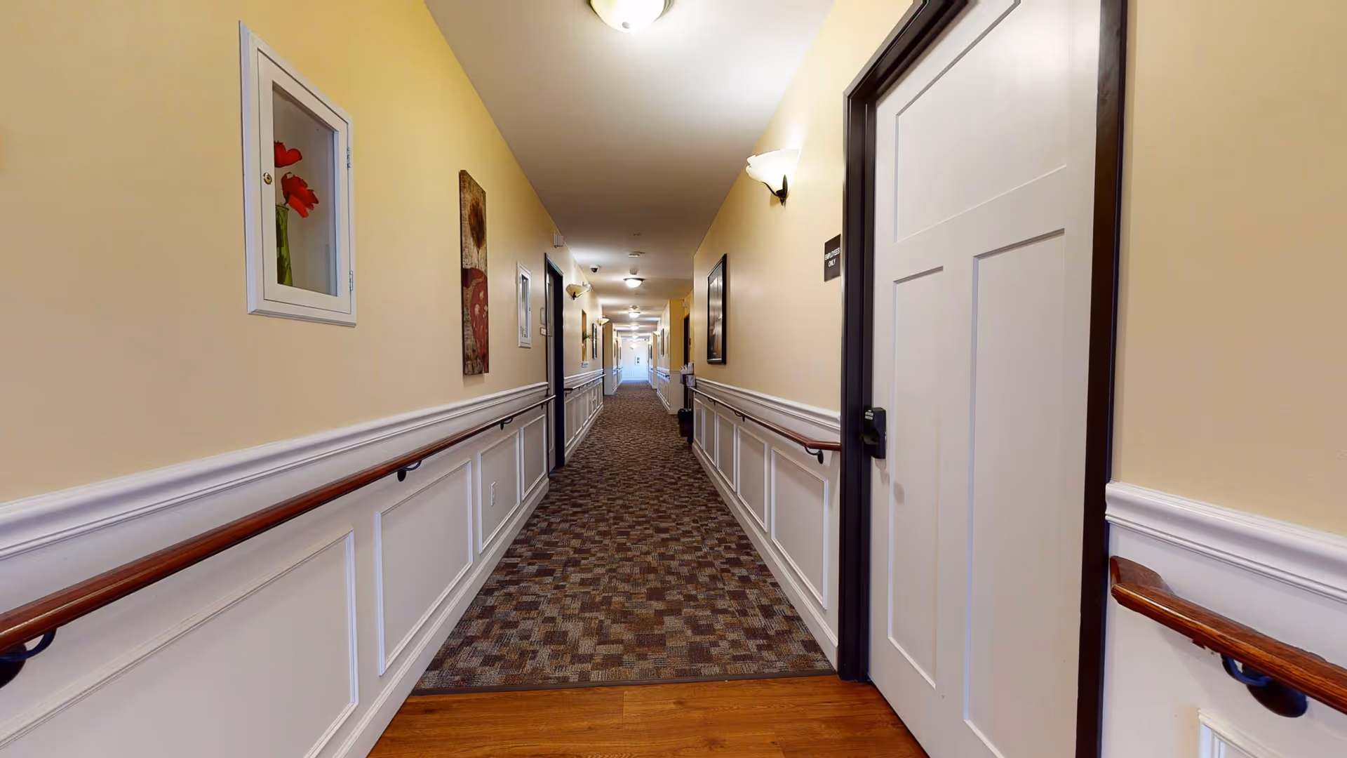 A long, well-lit hallway in a senior living facility with beige walls, white wainscoting, wooden handrails on both sides, and patterned carpet flooring. Several closed doors and framed artwork are visible along the walls, with ceiling lights evenly spaced along the corridor.