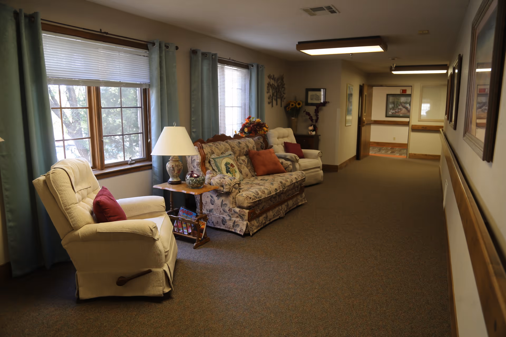 A cozy hallway seating area with two armchairs and a floral-patterned sofa, each adorned with colorful cushions. A wooden side table with a decorative lamp and magazines sits between the chairs. Large windows with teal curtains let in natural light, and framed artwork and floral decorations adorn the walls.