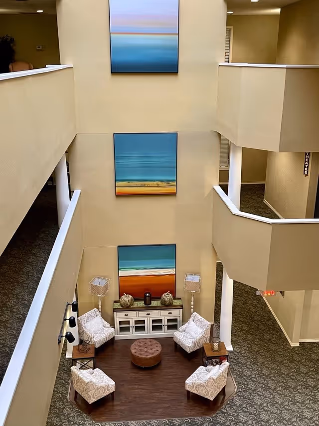View from above of a seating area in a multi-story interior space with four patterned armchairs arranged around a round brown ottoman on a wooden floor section. Behind the chairs is a white console table with decorative items and two tall lamps. Three large colorful abstract paintings are hung vertically on the beige wall. The surrounding floors have beige walls and patterned carpet.