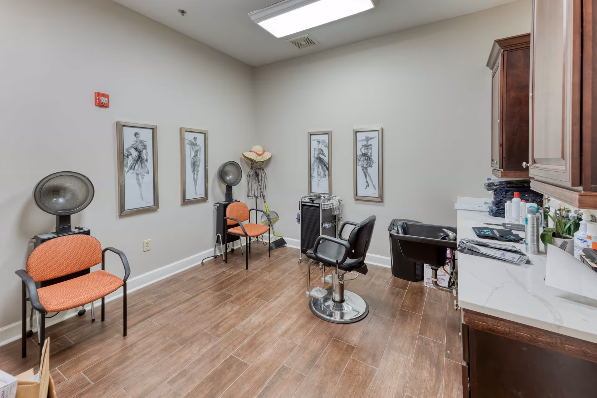 Small salon room with styling chairs, hooded hair dryers, a shampoo sink, framed artwork, and cabinets.