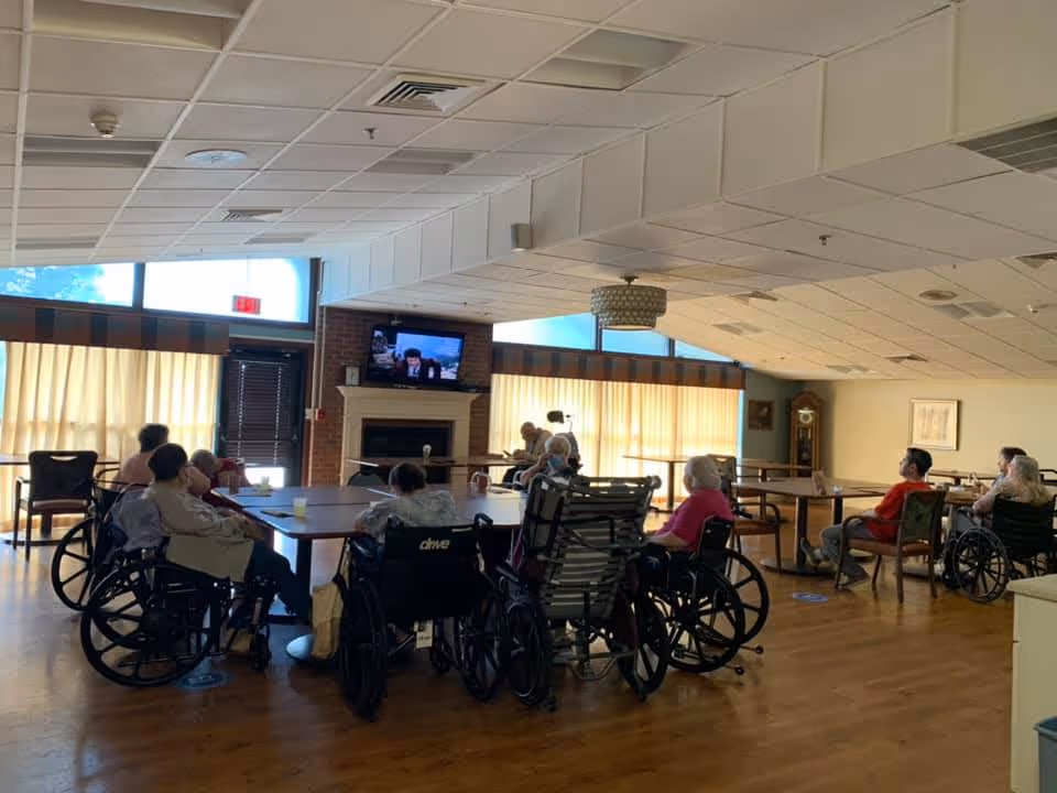 Residents in wheelchairs seated around tables in a senior facility common room watching a TV above a fireplace.