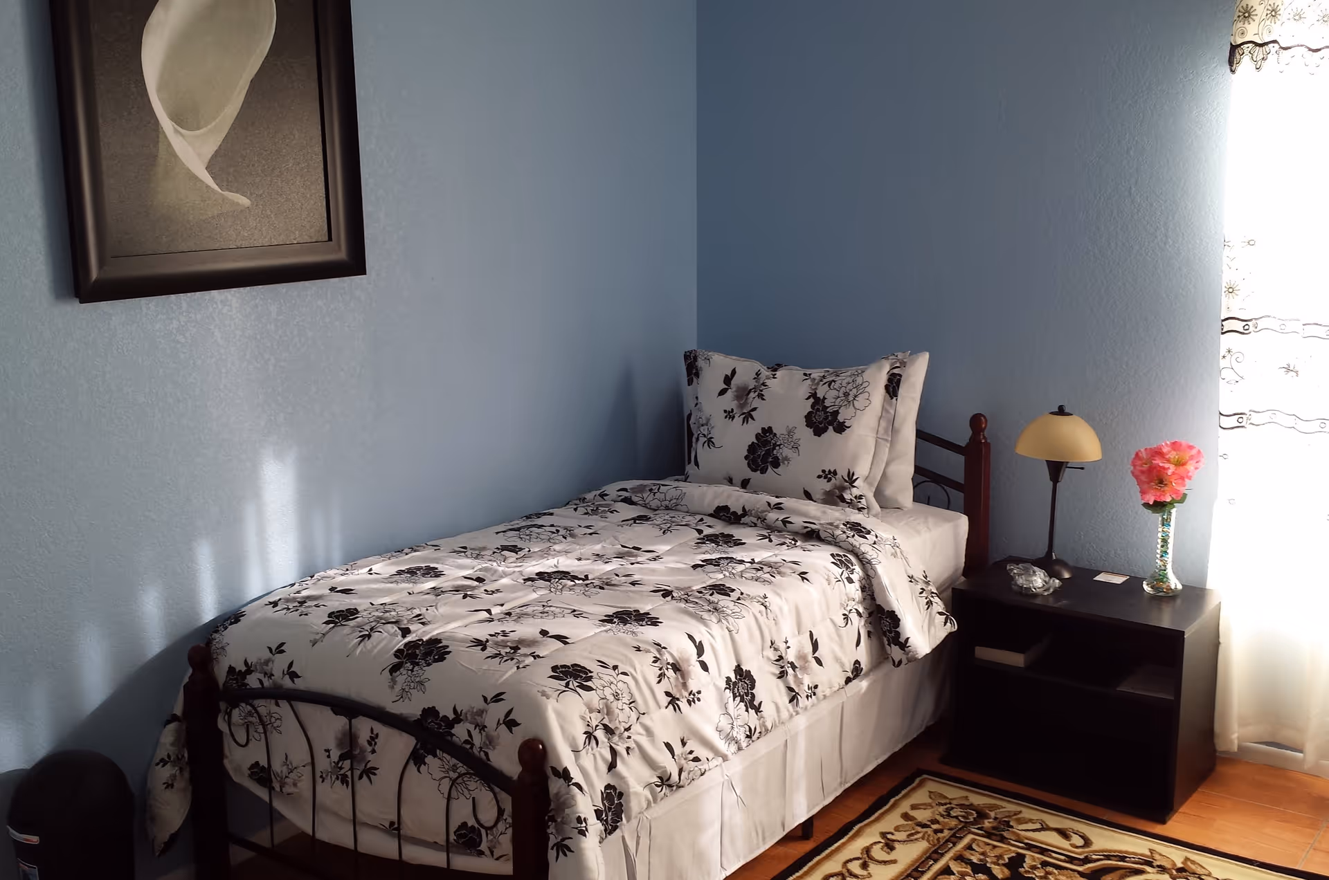 A small bedroom with light blue walls featuring a single bed with a black and white floral bedspread and pillow. Next to the bed is a dark wooden nightstand with a small lamp, a vase with pink flowers, and some small items. A framed black and white picture hangs on the wall above the bed, and a window with sheer curtains allows natural light to brighten the room. A patterned rug is partially visible on the floor.