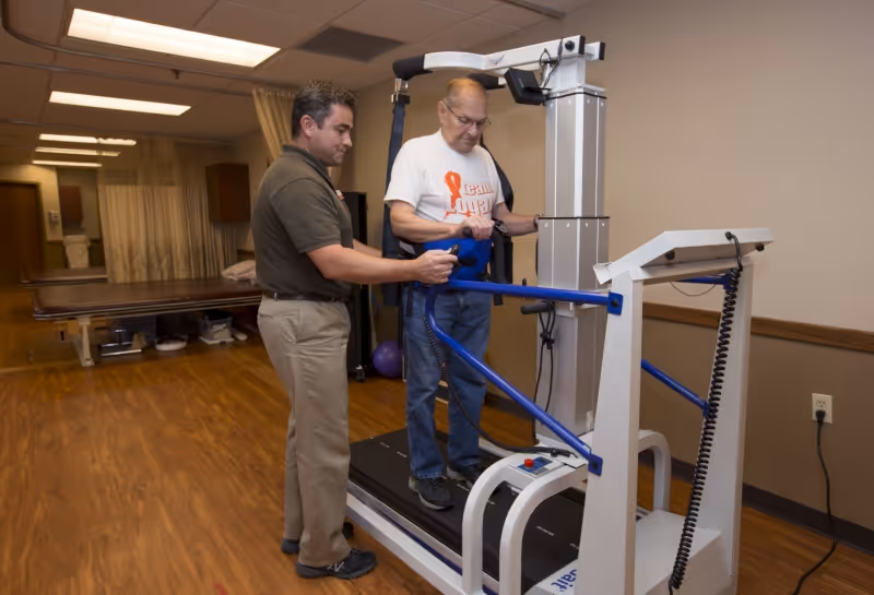A healthcare worker assists an elderly man using a treadmill with support bars in a rehabilitation room with wooden flooring and medical equipment in the background.