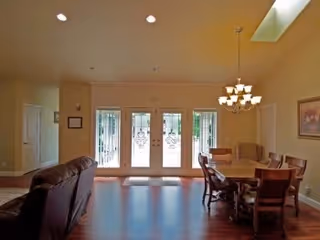 A spacious dining area with a wooden dining table surrounded by chairs and a built-in bench. The room has hardwood floors, a chandelier hanging from the ceiling, and large glass doors with decorative metalwork leading outside. The walls are painted a warm beige color, and there is a framed picture on one wall.