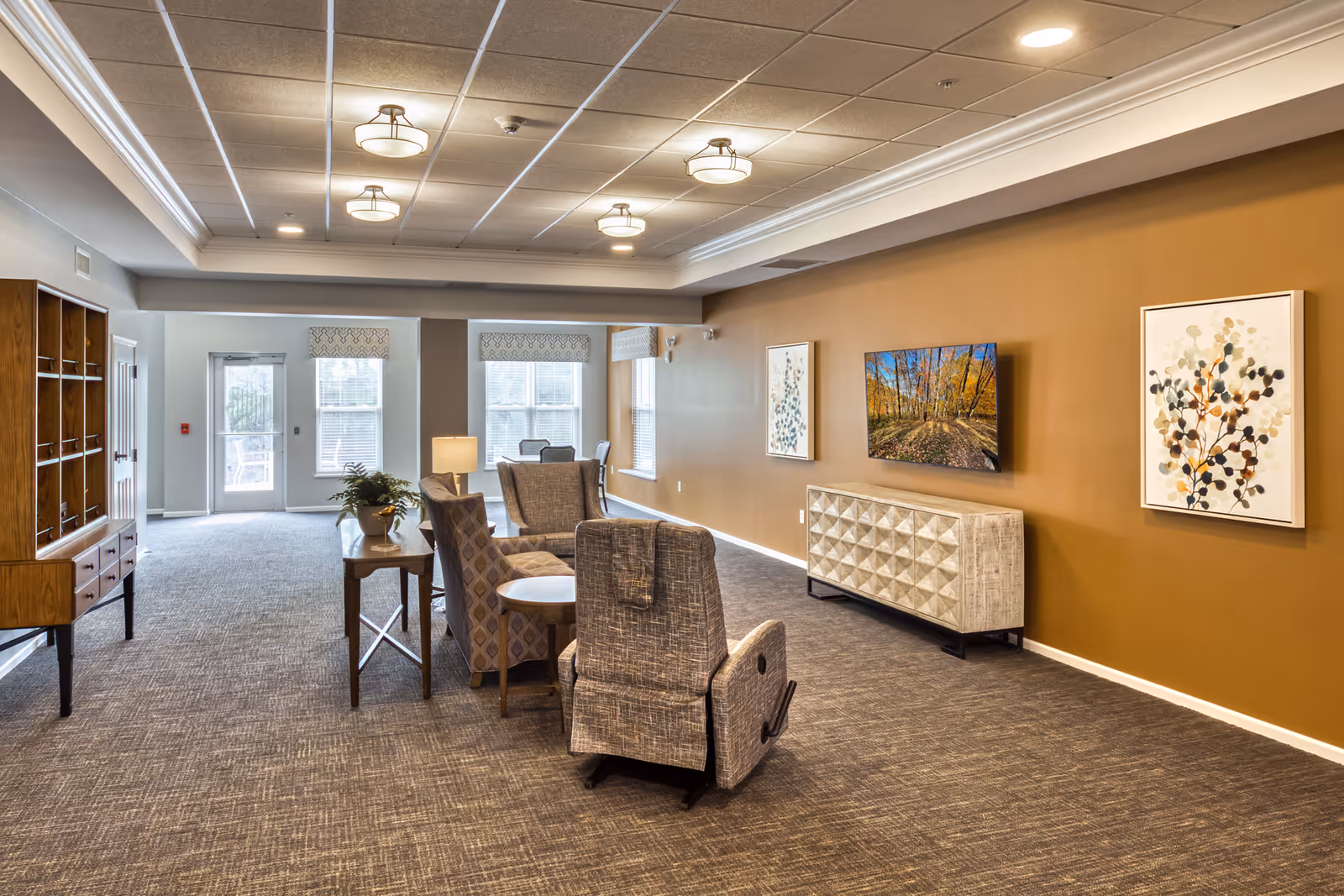 A spacious and well-lit common area in a senior living facility with comfortable upholstered chairs arranged around small tables. The room features a brown accent wall with two framed artworks and a mounted flat-screen TV above a decorative console. Large windows and a glass door allow natural light to fill the space, and ceiling lights provide additional illumination. A wooden shelving unit is visible on the left side of the room.