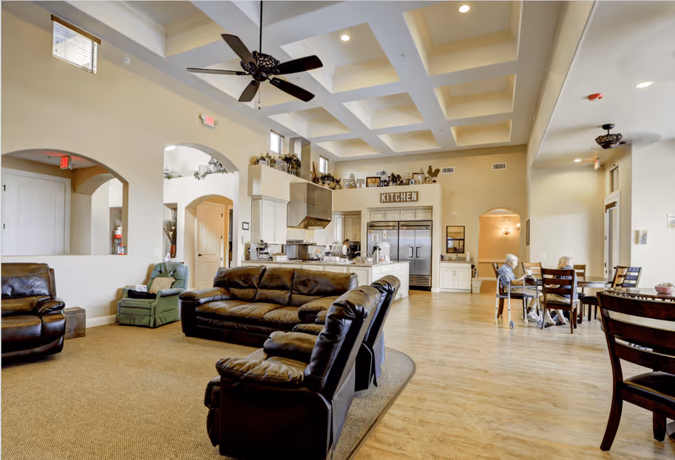 A spacious senior living common area featuring a seating area with leather sofas and chairs on a carpeted section, adjacent to a kitchen with white cabinetry and stainless steel appliances. Two elderly women are seated at a wooden dining table on the right side of the room. The ceiling has a coffered design with recessed lighting and ceiling fans. The room has large windows allowing natural light to fill the space.