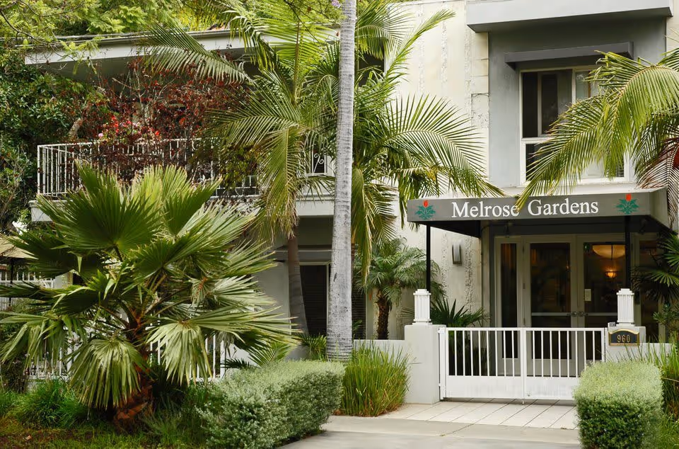 Entrance to Melrose Gardens facility surrounded by lush green palm trees and bushes, with a white gate and a canopy displaying the name Melrose Gardens.