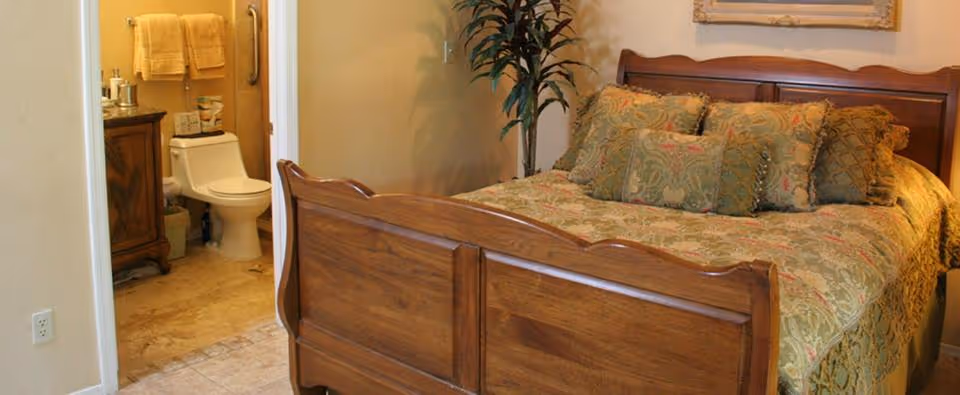Bedroom with a wooden bed and patterned bedspread, and an open doorway revealing a bathroom with a toilet and vanity.