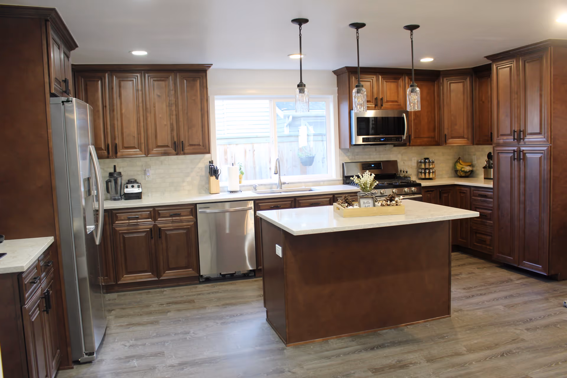 A spacious modern kitchen with dark wood cabinets, a central island with a white countertop, stainless steel appliances, and pendant lights.